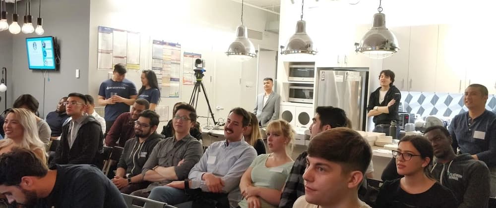 A group of people attentively watch a presentation in a modern, well-lit room with industrial pendant lights, a kitchen area, and a mounted TV screen. The atmosphere is engaged and focused.